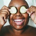 Black woman smiling holding cucumber slices over her eyes hair up in towel