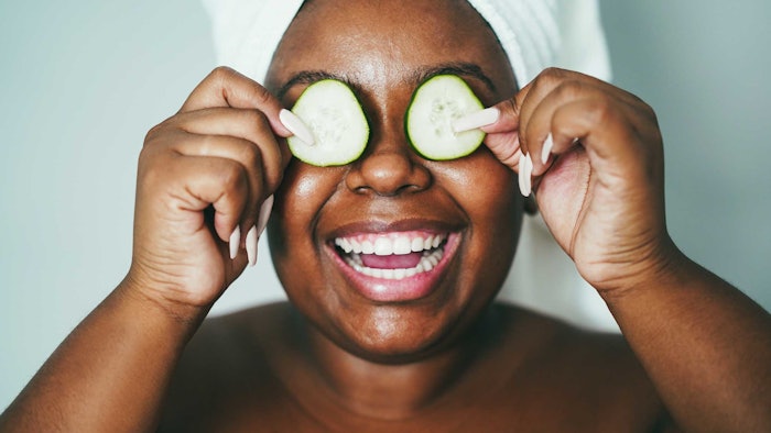 Black woman smiling holding cucumber slices over her eyes hair up in towel