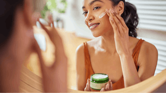 medium-toned woman applying natural skin cream in mirror smiling