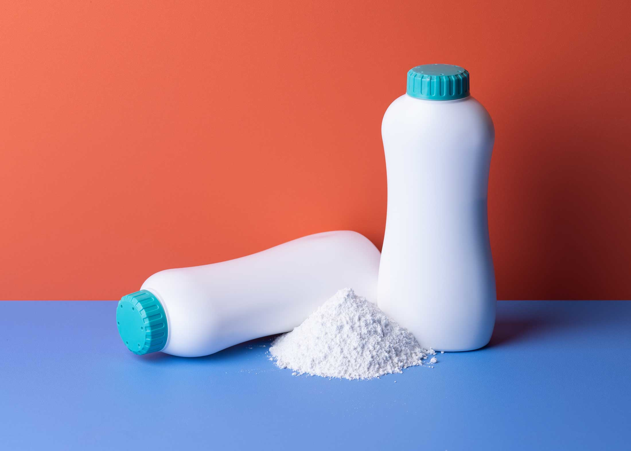 two bottles of talc powder one spilled over blue surface red background AdobeStock_687808176
