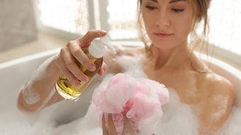 woman in foamy bath pouring body gel into loofah