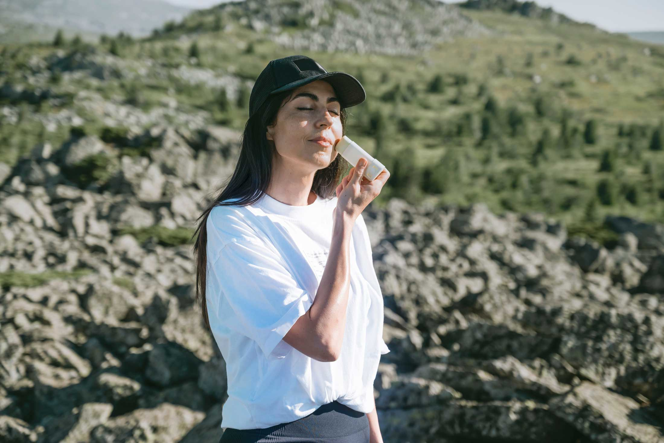 woman in mountains wearing hat using stick sunscreen on face