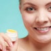 young caucasian woman smiling dipped finger into jar of balm teal bg