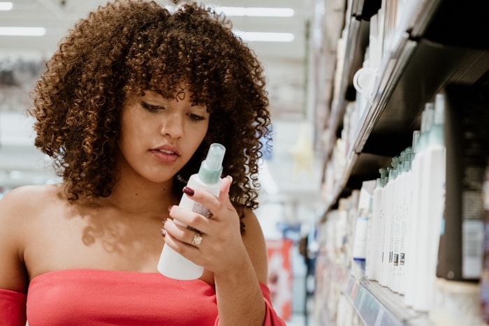 A woman with curly hair holds a hair product in her hand while standing in a hair care aisle in a supermarket.