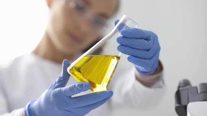 female chemist in lab coat with gloves examining gold liquid in flask