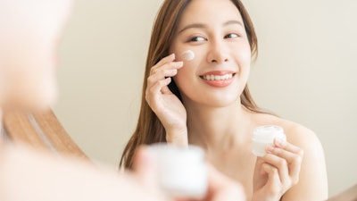 beautiful Asian woman in mirror applying skin cream to face holding jar