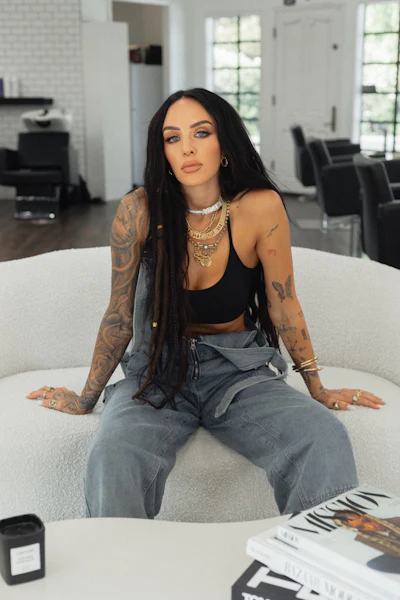 Tattooed woman in black top and baggy jeans sitting on white chair in modern interior