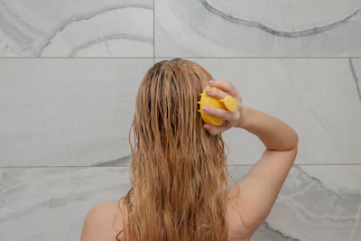 Person applying yellow shower sponge to wet hair in bathroom with marble tile background