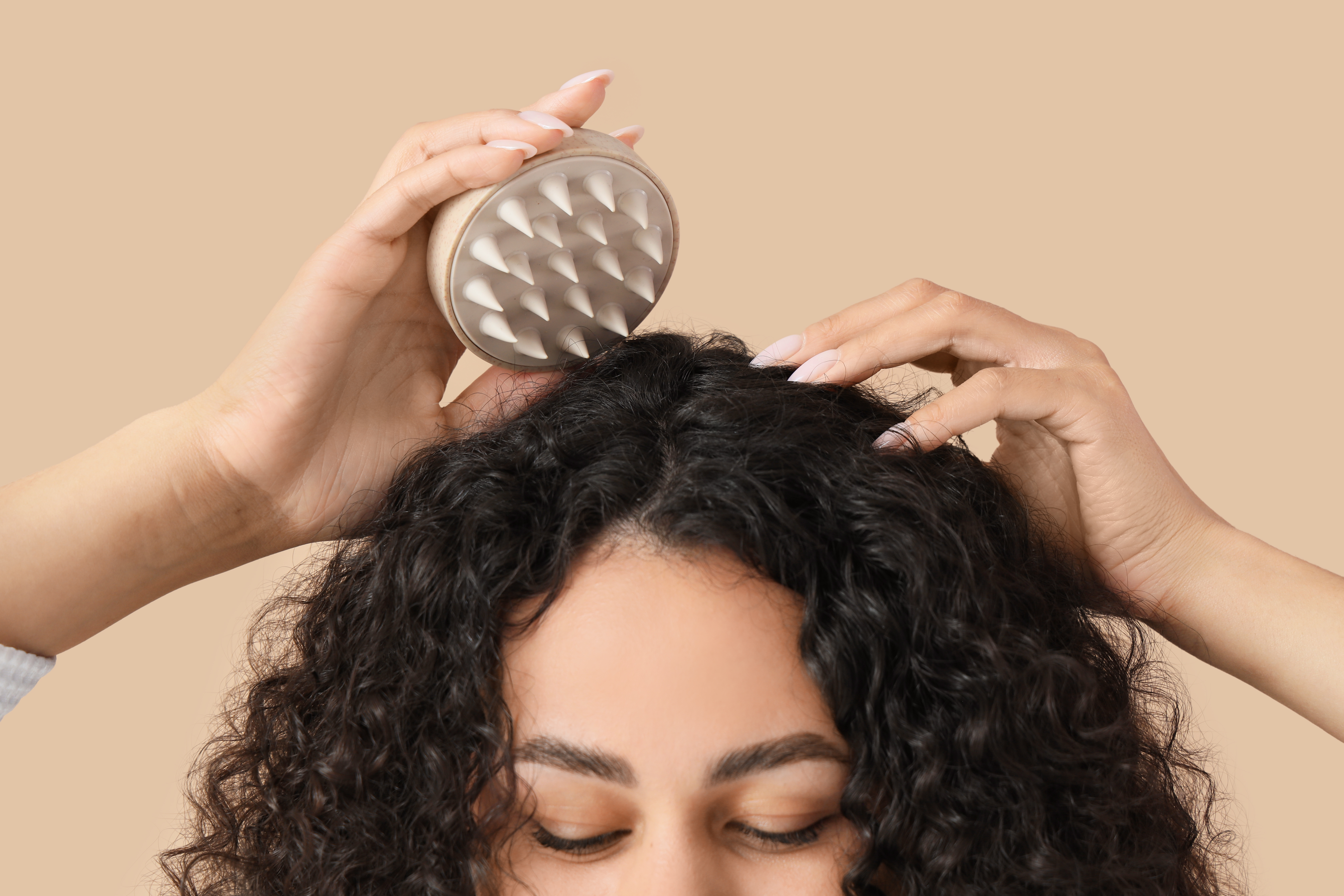 Person using a metal scalp massager on curly black hair against a neutral beige background