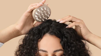 Person using a metal scalp massager on curly black hair against a neutral beige background