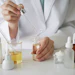Scientist in white lab coat pipetting golden serum into glass bottle surrounded by skincare products and formulations
