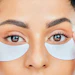 Woman applying white under-eye hydrating patches against a blue background