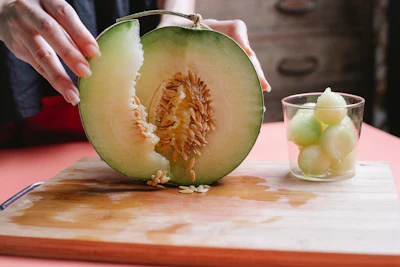 Hands holding halved cantaloupe melon with seeds visible, melon balls in glass on cutting board