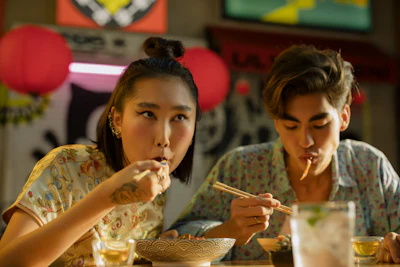 Two people eating noodles with chopsticks at a table in a casual dining setting with colorful balloons in the background