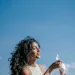 Woman in cream tank top holding beverage bottles outdoors against clear blue sky