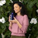 Woman in mauve sweater holding blue cosmetic bottle against green foliage and white flowers