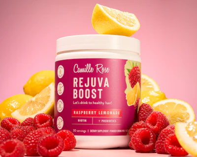 Camille Rose Rejuva Boost dietary supplement jar with raspberry lemonade flavor surrounded by fresh raspberries and lemon slices on pink background