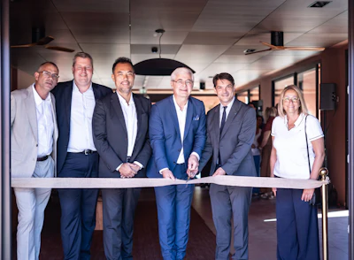 Six business professionals in formal attire participate in a ribbon-cutting ceremony in a modern office building.