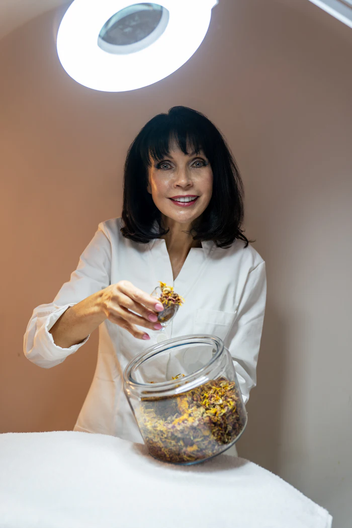 Woman in white lab coat holding dried herbs from glass jar under ring light in professional setting