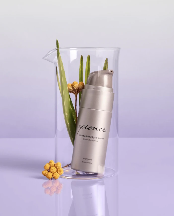 Epionce Ultra Refining Lytic Serum bottle displayed in clear cylinder with green plant stems and yellow flowers on lavender background