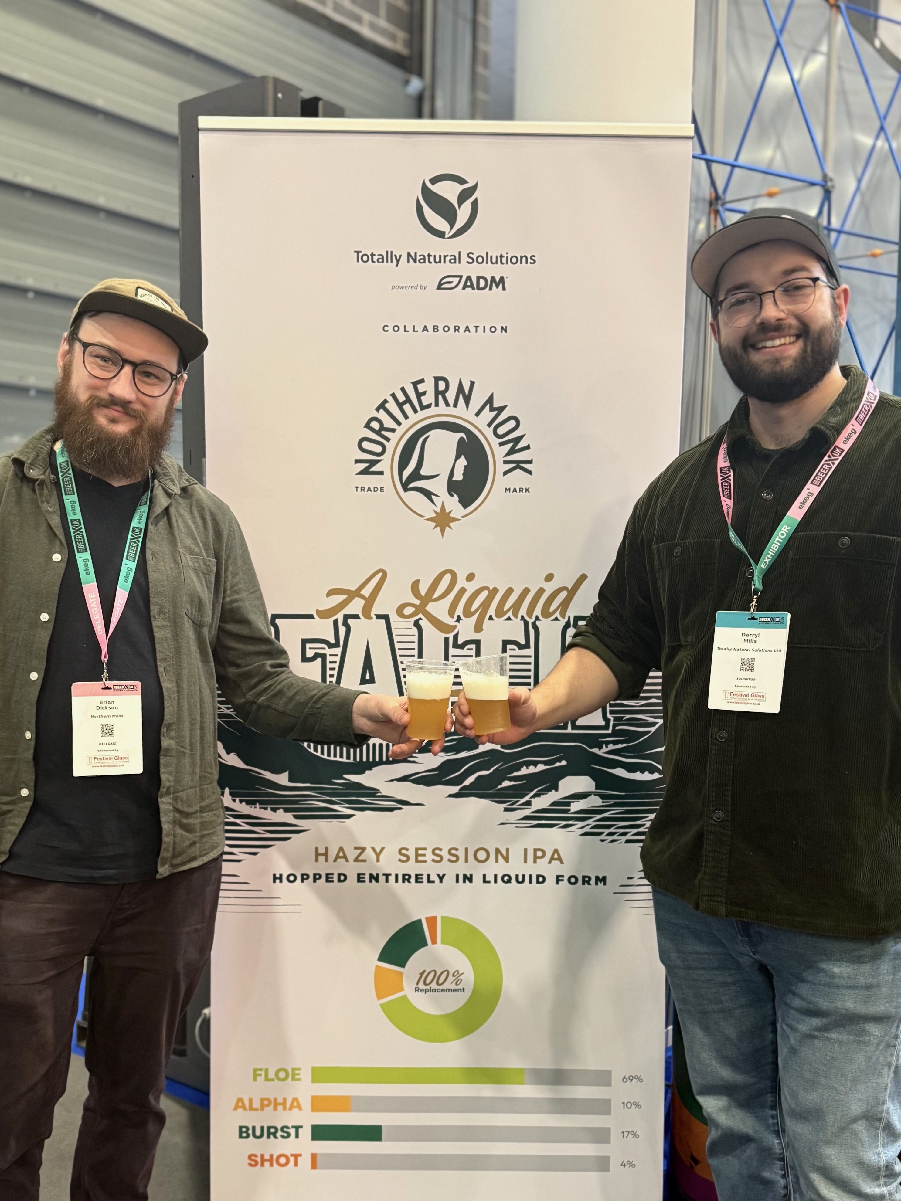 Two men with event badges stand beside a Northern Monk brewery collaboration poster for A Liquid Fate Hazy Session IPA beer