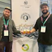 Two men with event badges stand beside a Northern Monk brewery collaboration poster for A Liquid Fate Hazy Session IPA beer