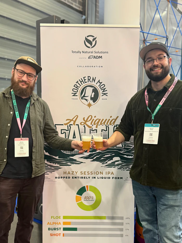 Two men with event badges stand beside a Northern Monk brewery collaboration poster for A Liquid Fate Hazy Session IPA beer