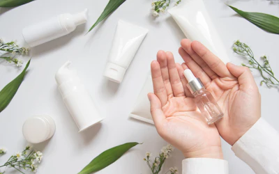 Hand holding clear serum bottle surrounded by white skincare products and green botanical leaves on white background