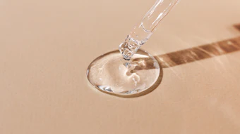 Clear liquid serum dispensed from dropper into glass dish on beige background