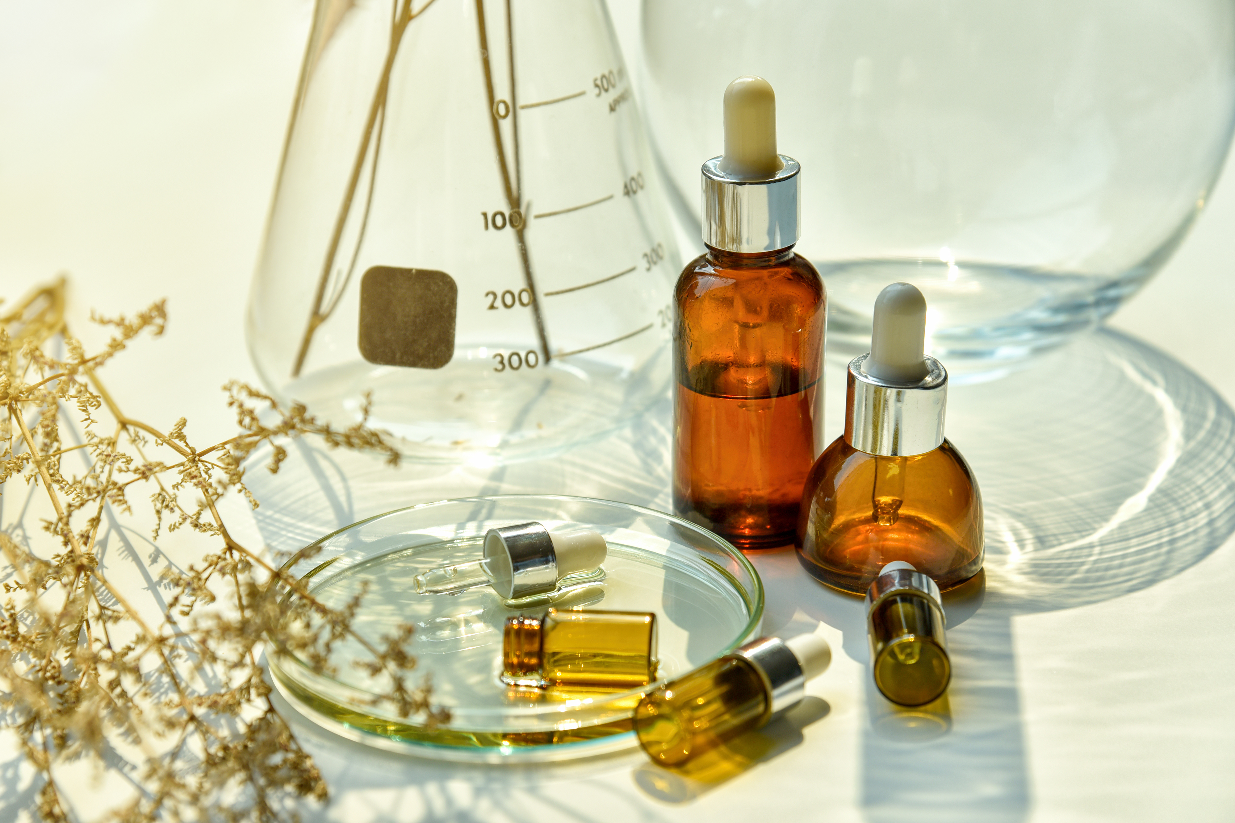 Amber glass dropper bottles with essential oils or serums arranged with laboratory beakers, dried flowers, and measurement scales on white surface