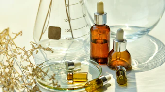 Amber glass dropper bottles with essential oils or serums arranged with laboratory beakers, dried flowers, and measurement scales on white surface
