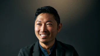 Professional headshot of an Asian man smiling at camera, wearing black blazer and dark blue shirt against dark background