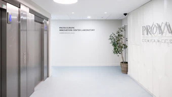 Modern office elevator lobby with PROVA branding, featuring stainless steel elevators, white walls, potted plant, and recessed ceiling lights