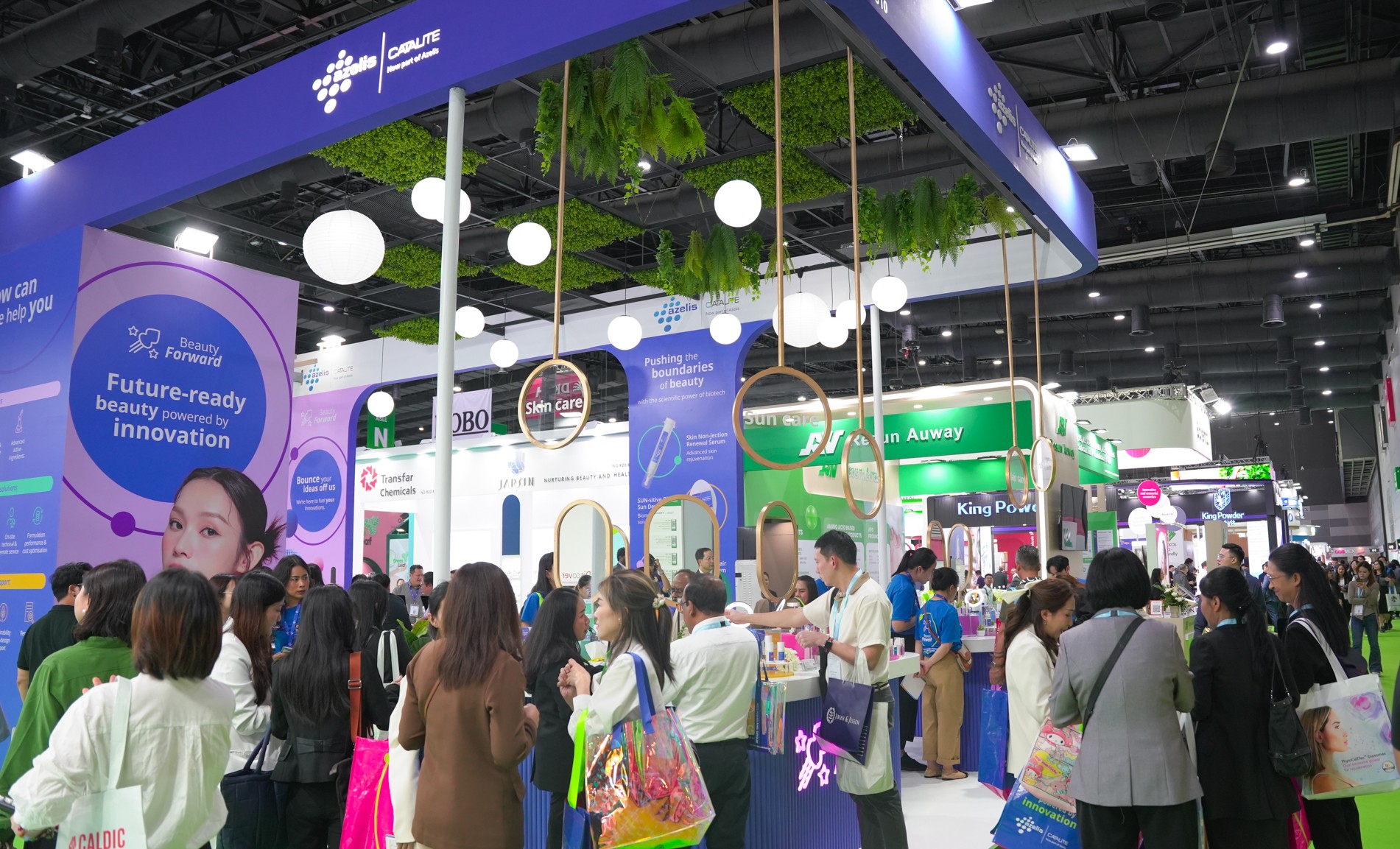 Crowded trade show booth with purple and blue branding, hanging lights, green plants, and visitors exploring displays