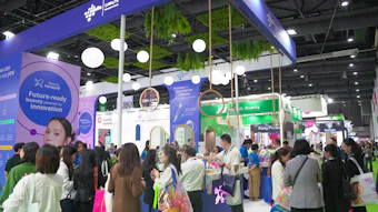 Crowded trade show booth with purple and blue branding, hanging lights, green plants, and visitors exploring displays