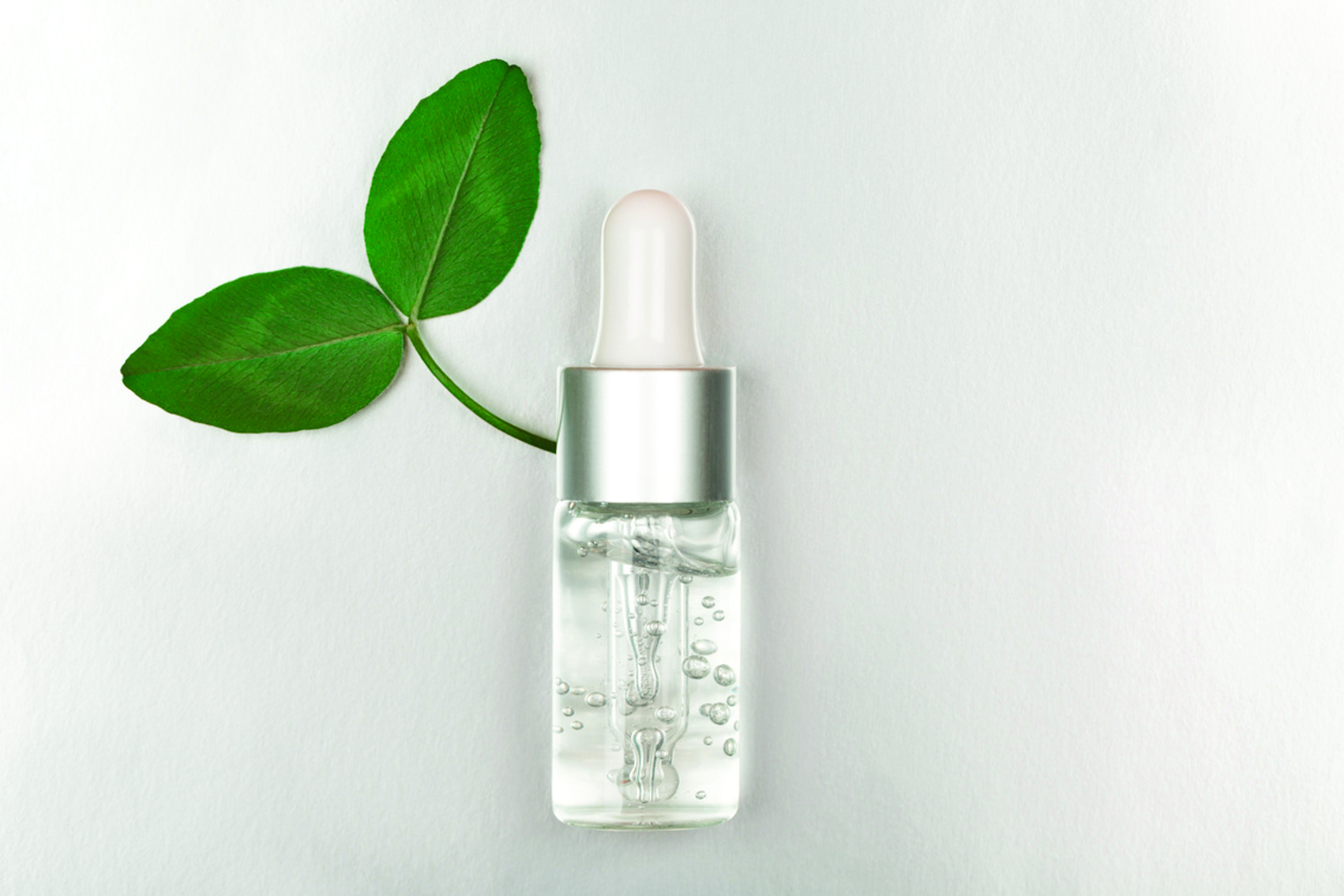 Clear serum bottle with silver dropper cap next to fresh green leaves on white background