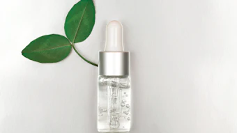 Clear serum bottle with silver dropper cap next to fresh green leaves on white background
