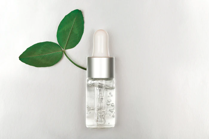 Clear serum bottle with silver dropper cap next to fresh green leaves on white background