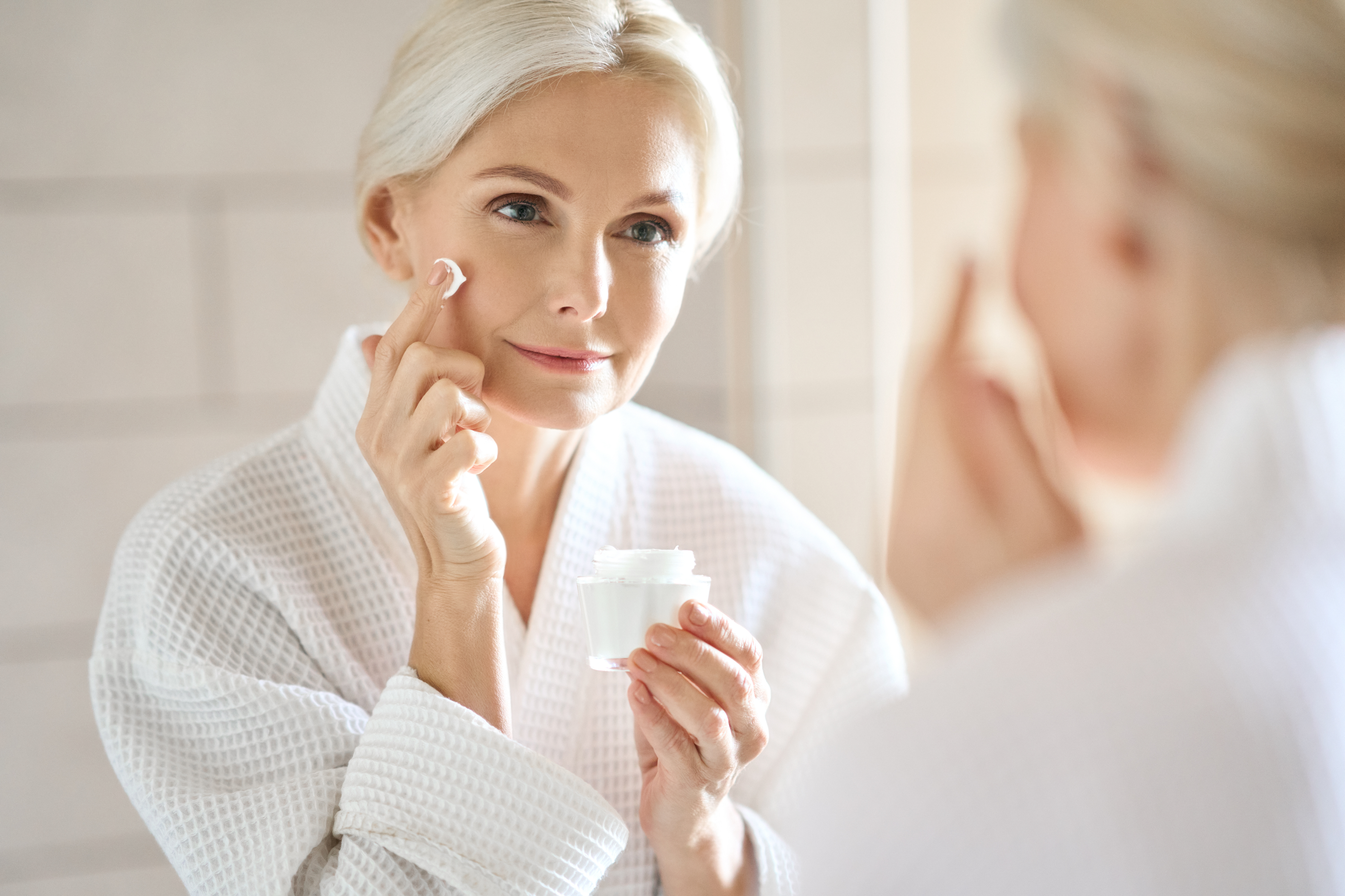 Mature woman applying facial cream while looking in mirror with confident expression