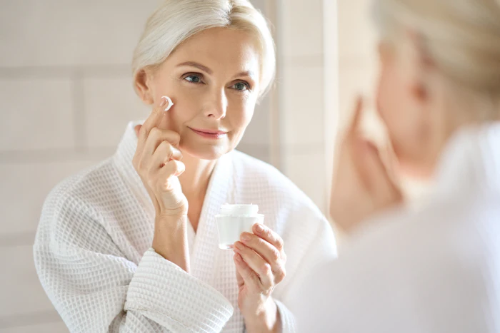 Mature woman applying facial cream while looking in mirror with confident expression