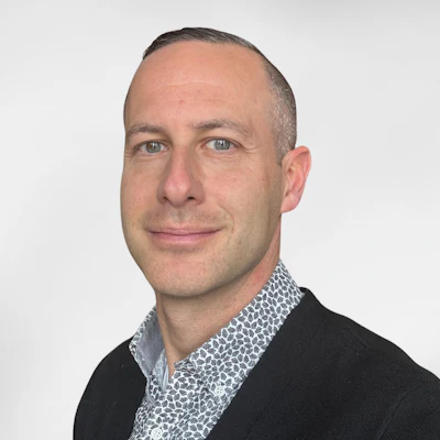 Professional headshot of a man wearing a black cardigan over a patterned shirt against a light gray background