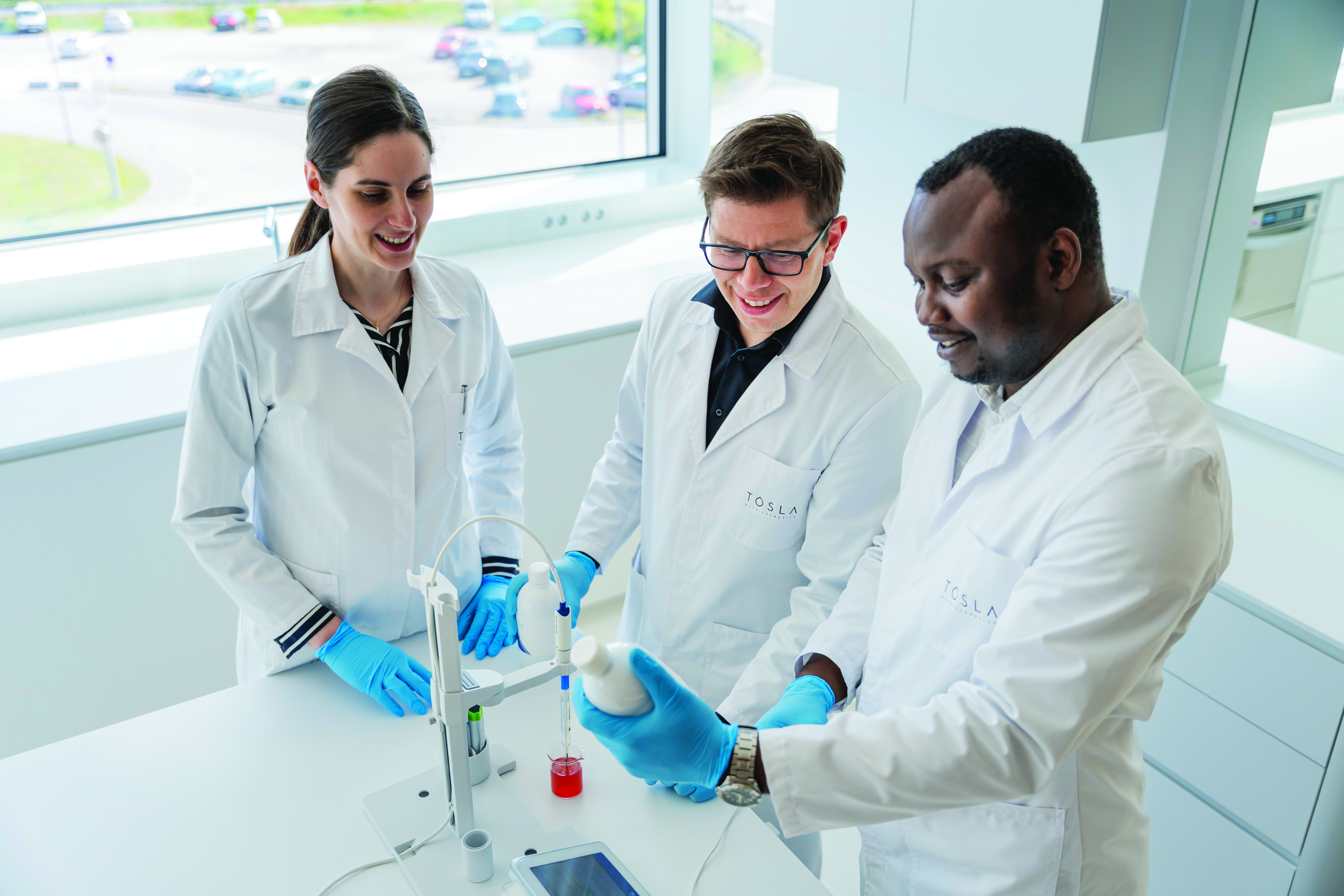 Three scientists in white lab coats conducting research with laboratory equipment and samples in a modern laboratory setting with large windows.